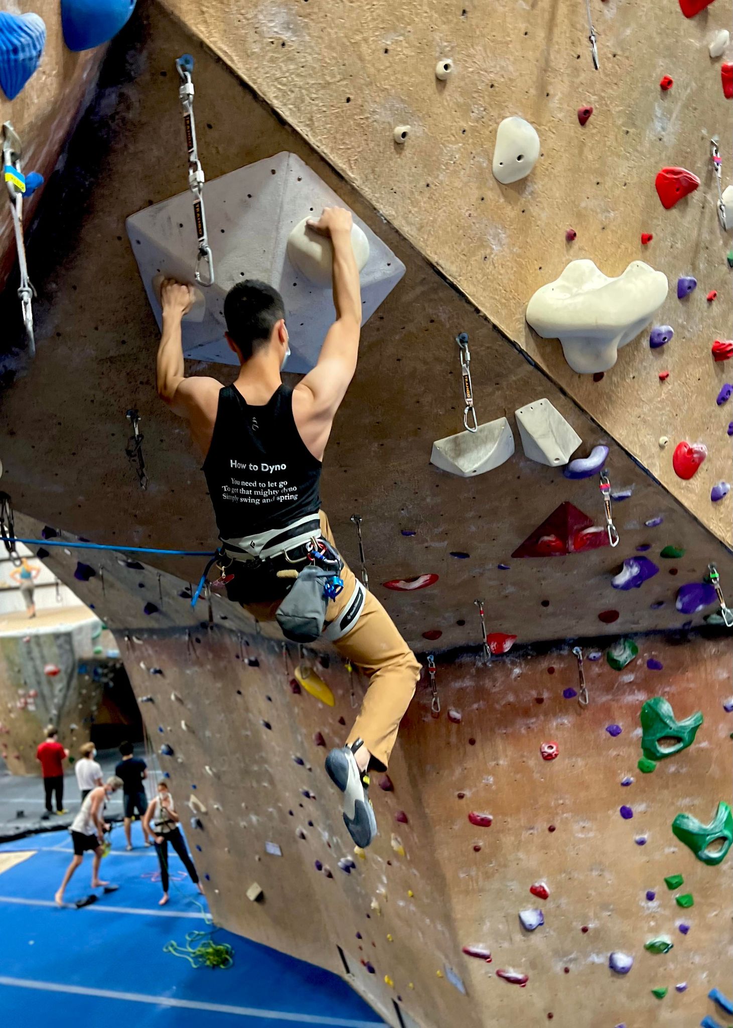 Charcoal black tank top shirt with How to Dyno haiku on the back worn by male lead climbing on a climbing wall in the gym