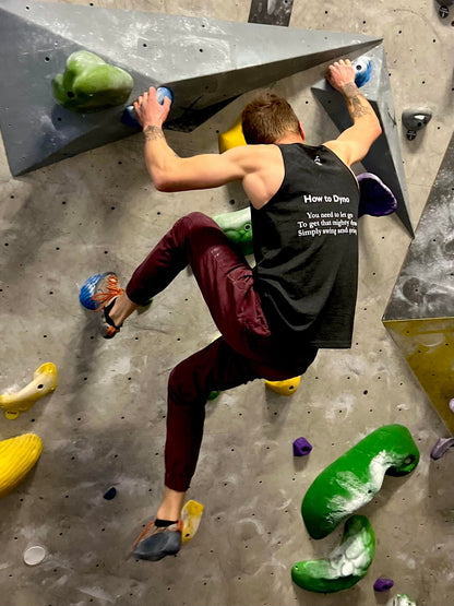Charcoal black tank top shirt with How to Dyno haiku on the back worn by male bouldering on a climbing wall in the gym