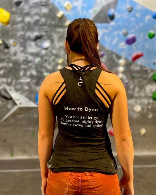 Charcoal black tank top shirt with How to Dyno haiku on the back worn by female looking at climbing wall in the gym