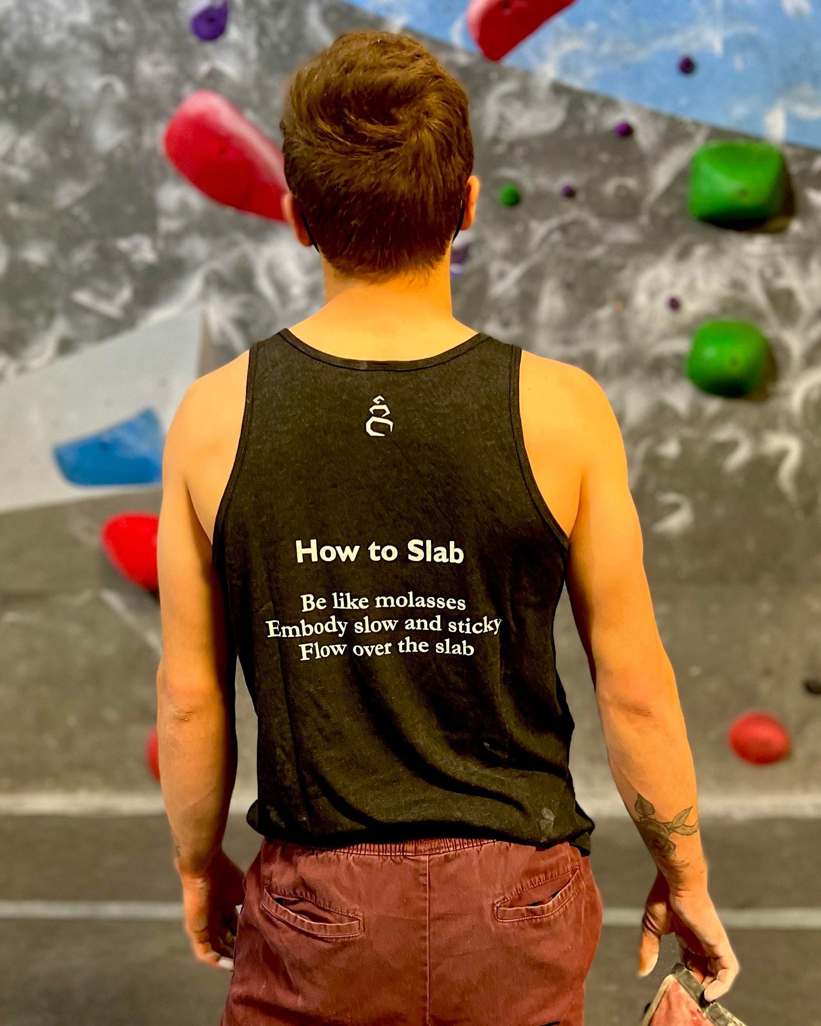 Charcoal black tank top shirt with How to Slab haiku on the back worn by male looking at climbing wall in the gym