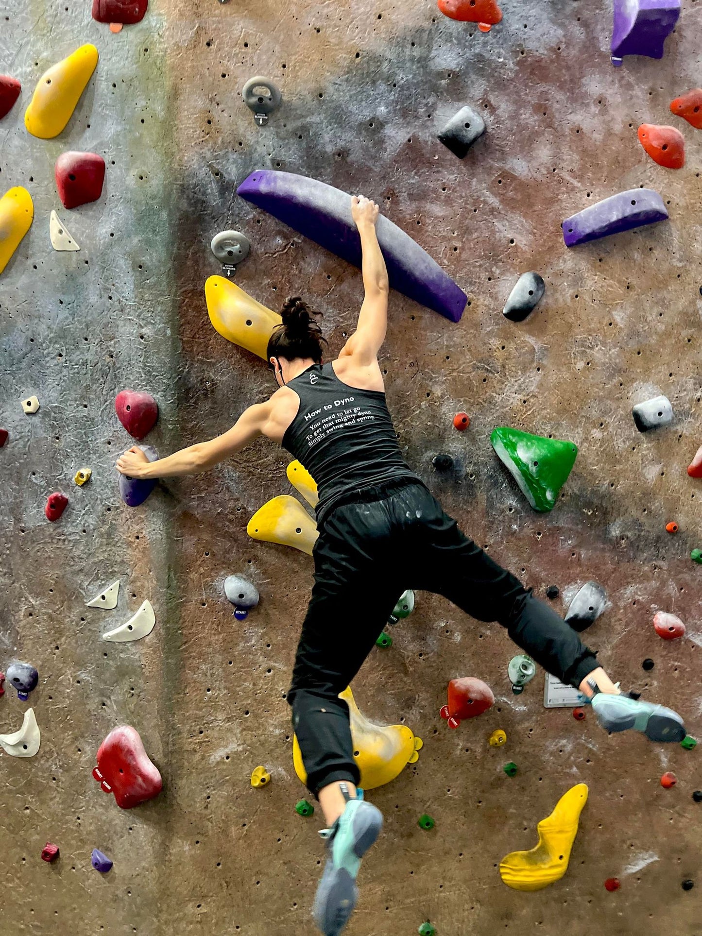 Charcoal black tank top shirt with How to Dyno haiku on the back worn by female bouldering on a climbing wall in the gym
