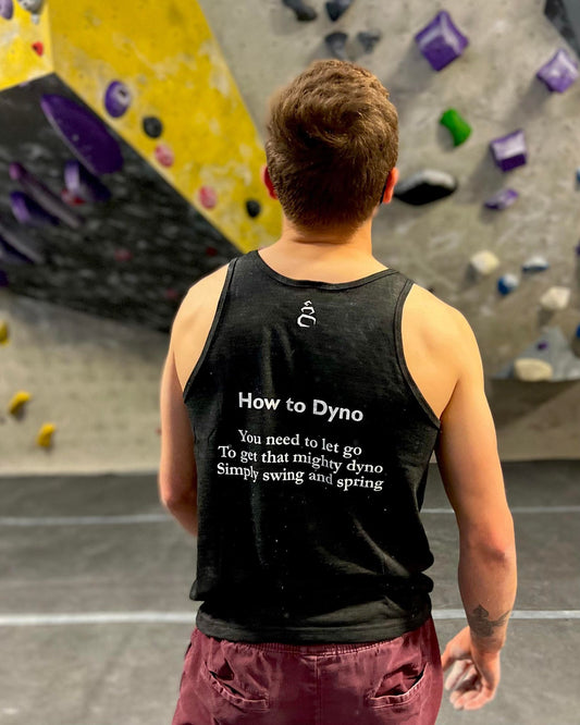 Charcoal black tank top shirt with How to Dyno haiku on the back worn by male looking at climbing wall in the gym